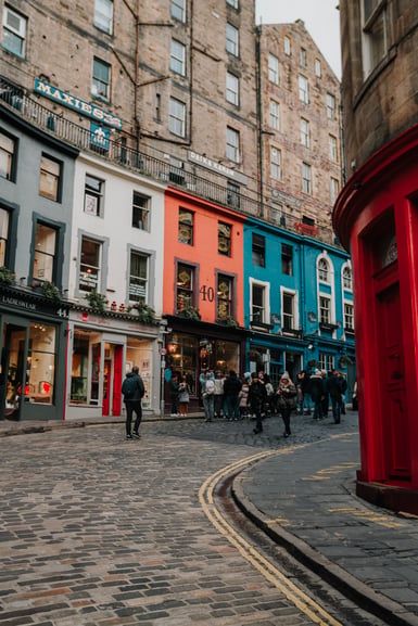 A curving cobbled street in Edinburgh lined with painted shopfronts in blue, white, orange and teal beneath tall stone tenements, with people walking near a red corner building