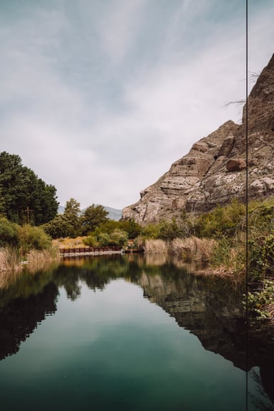 A still green pond edged with reeds and trees reflects a pale layered rock cliff on the right under an overcast sky, with a wooden footbridge across the far end and a thin vertical cable crossing the right side of the frame