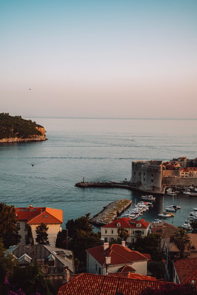 Elevated view of a small harbour with moored boats beside a round stone fortress tower, terracotta rooftops in the foreground and a calm sea meeting a pale sky