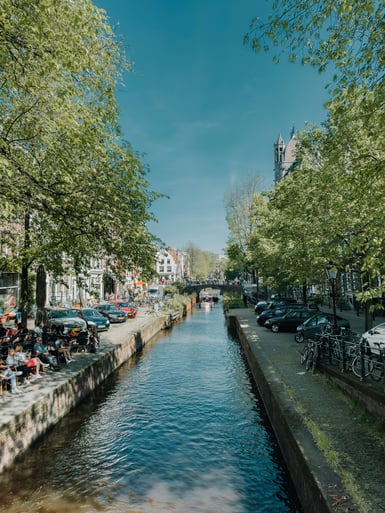 A narrow Amsterdam canal in spring, flanked by elms, parked cars and bicycles, with people sitting at a waterside café on the left and a church tower behind