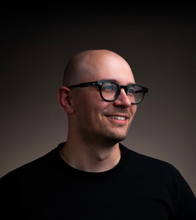Studio portrait of a smiling bald man with a short beard, wearing black round-framed glasses and a black t-shirt, looking off to his left against a dark brown backdrop