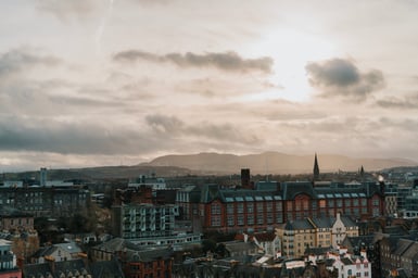 Elevated view over Edinburgh rooftops and a large red sandstone building with a church spire, with low hills on the horizon under a cloudy, hazy sky