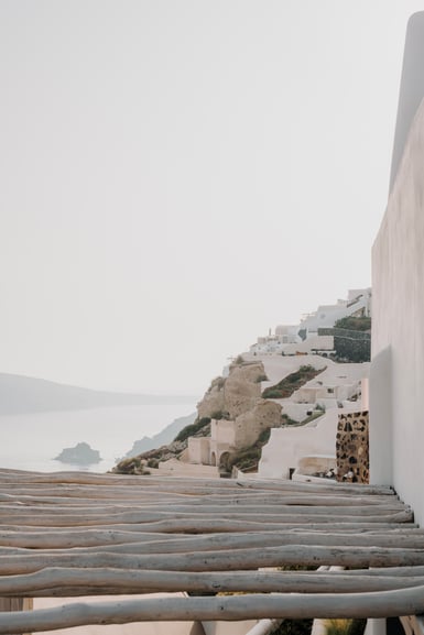 Whitewashed buildings step down a rocky cliff on the right above a pale hazy sea, with bare wooden pergola poles crossing the lower foreground