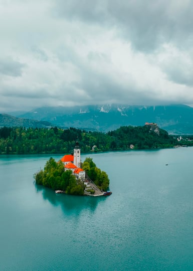 A small wooded island with a red-roofed church and bell tower sits in turquoise Lake Bled, with forested hills and low cloud behind