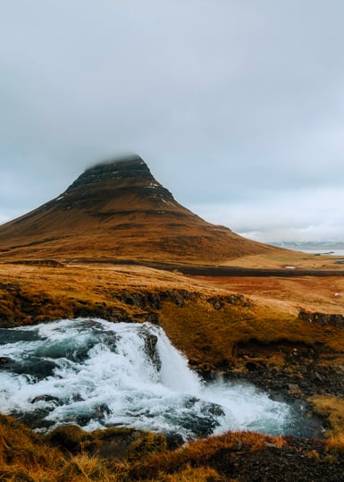 A steep isolated mountain with its peak hidden in low grey cloud rises above brown moorland, with a small white waterfall spilling over dark rock in the foreground