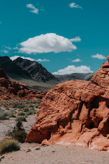 A red sandstone outcrop pitted with small holes rises at right above pale desert gravel, with dark grey mountains and a single white cloud behind