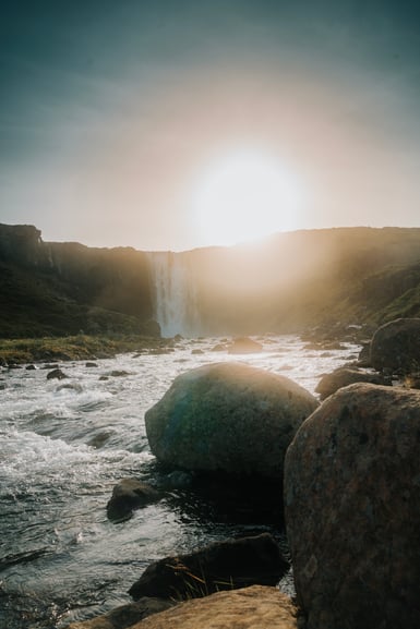 A waterfall drops from a low cliff into a rocky river in east Iceland, with the sun low and hazy directly above it and large rounded boulders in the foreground