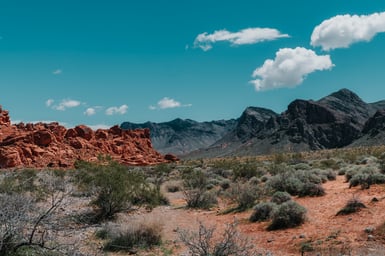 A desert basin with low red sandstone outcrops on the left, grey rocky mountains along the horizon, scattered pale shrubs on orange sandy ground, and white clouds in a deep blue sky