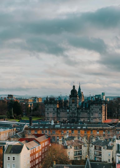 Elevated view over Edinburgh rooftops and stone buildings with domed towers, a pale strip of water and low hills on the horizon under heavy grey cloud