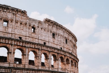 Close view of the Colosseum's curving upper wall in Rome, with a row of arched openings below, rectangular holes above, and pale sky with light cloud behind