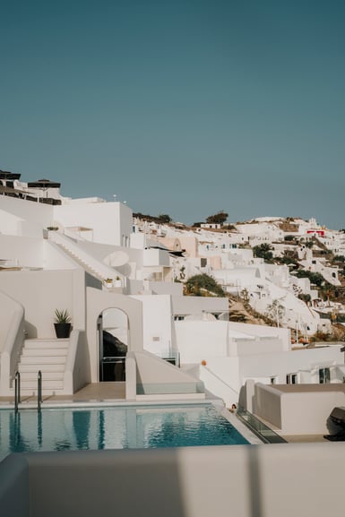 Whitewashed Cycladic buildings stacked up a hillside under a clear blue sky, with a small swimming pool, white stairs and a potted plant in the foreground