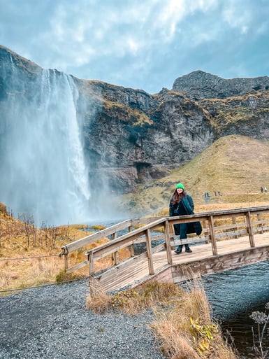 A woman in a green knitted hat and dark coat stands on a wooden footbridge over a stream, with a tall waterfall spilling off a cliff behind her