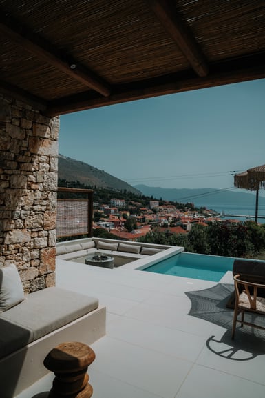 A shaded terrace with reed-covered pergola, stone wall and built-in cushioned bench looks out over a small pool toward a red-roofed hillside village and blue sea