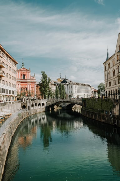 A calm green river runs between stone embankments in Ljubljana, with a triple arched bridge, a pink baroque church and pale townhouses reflected under a cloudy sky