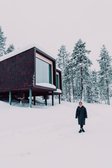 A dark shingled cabin raised on thin stilts above deep snow, with large windows facing snow-covered pines, and a person in a long dark coat standing in the snow at right