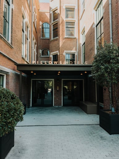 A brick building courtyard leading to a dark glass hotel entrance under a black canopy lettered "Pillows Hotels", with clipped shrubs and a small tree in black planters flanking the paved walkway