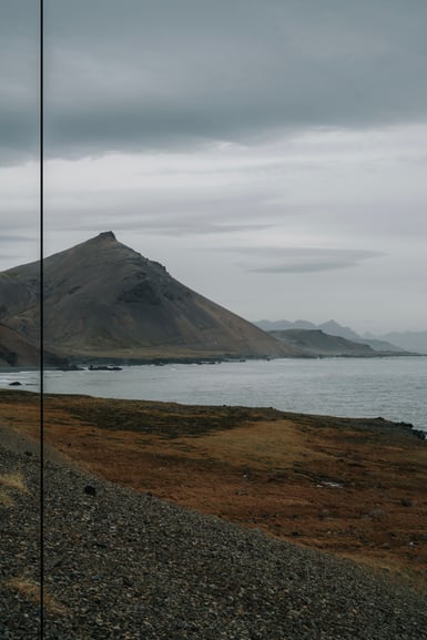 A conical brown mountain rises above a grey coastline under heavy overcast cloud, with gravel and rust-coloured grass in the foreground and a thin vertical line crossing the left side of the frame