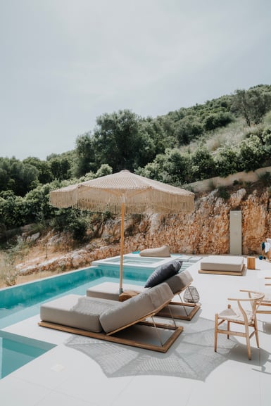 A swimming pool terrace with two beige loungers, a fringed umbrella and wicker chairs, backed by a cut limestone bank and scrub-covered hillside under a pale sky