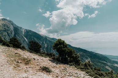 A stony white slope with scattered pines in the foreground drops away toward a limestone ridge and a hazy sea horizon under tall cumulus cloud