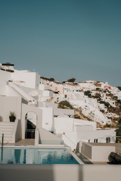Whitewashed Cycladic buildings with staircases and arched doorways climbing a dry hillside under a clear blue sky, with the edge of a swimming pool in the foreground