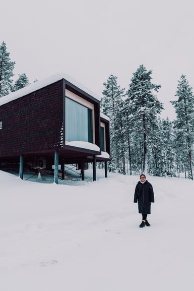 A person in a long dark coat stands on deep snow in front of a dark timber cabin raised on slender stilts, with snow-covered spruce and pine trees behind under a white sky