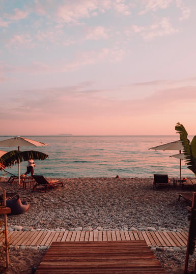 A pebble beach at dusk with two closed white parasols, sun loungers and a wooden boardwalk leading toward a calm sea under a pink sky