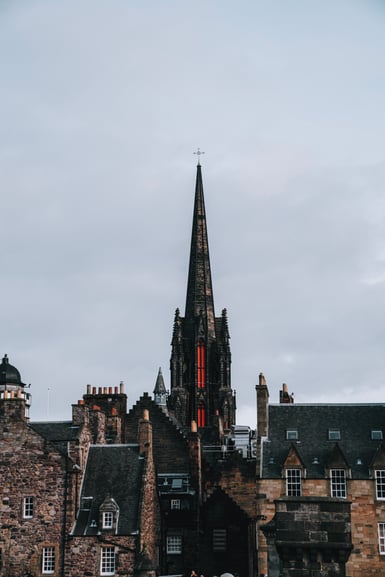 A tall dark Gothic church spire topped with a cross rises above stone tenement rooftops and chimney stacks under a flat grey overcast sky, with orange netting visible on the spire's lower section